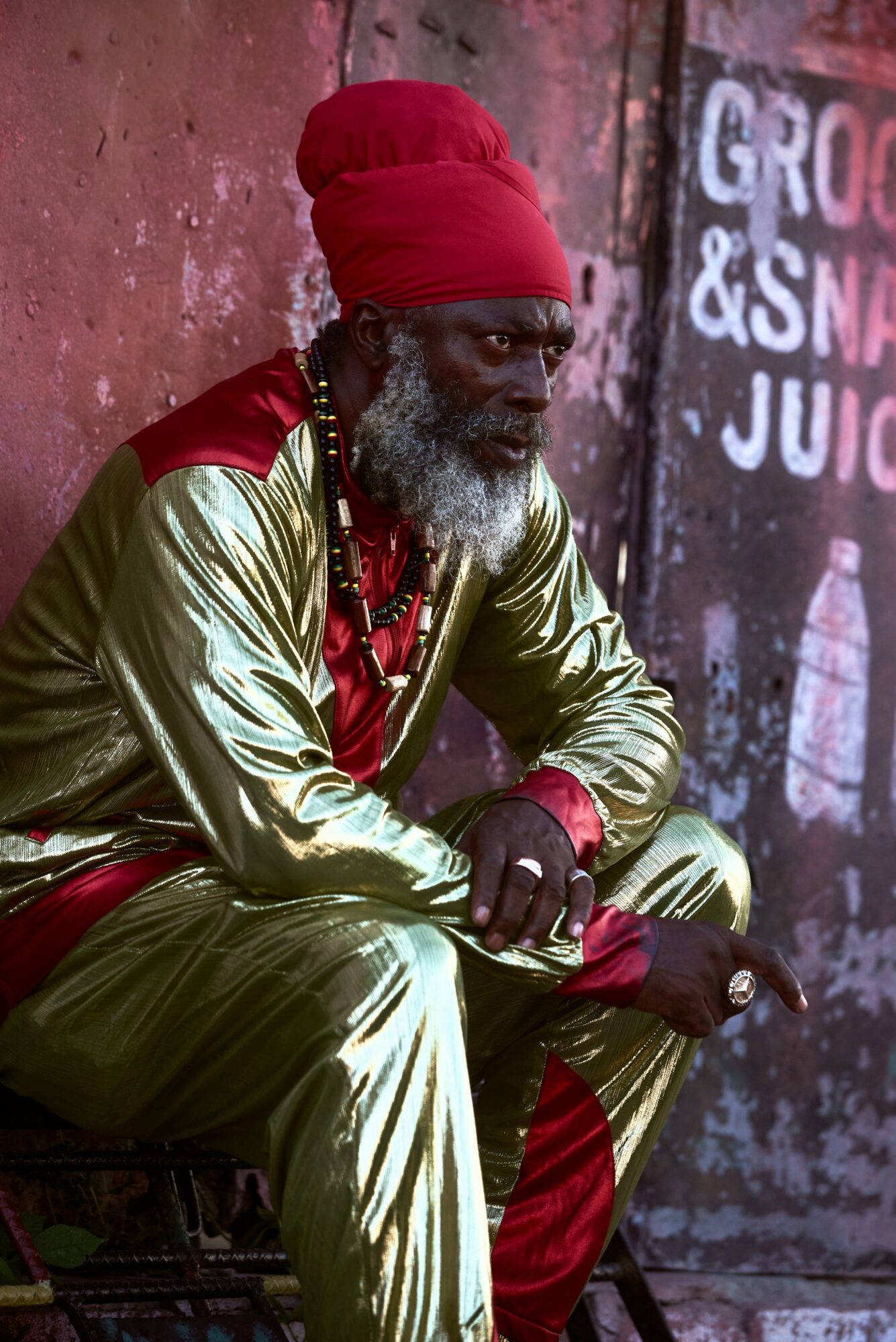 Elderly man with a gray beard wearing a red headwrap and shiny gold clothing, sitting outdoors near a wall with graffiti.