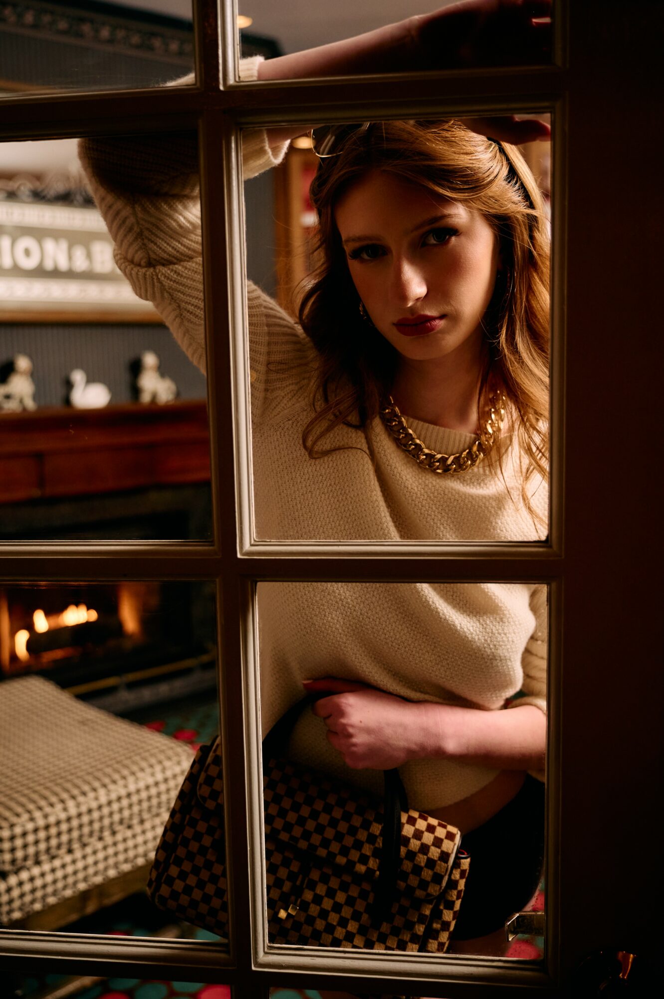 Young woman with wavy hair and a necklace looks through a window, indoors with warm lighting.