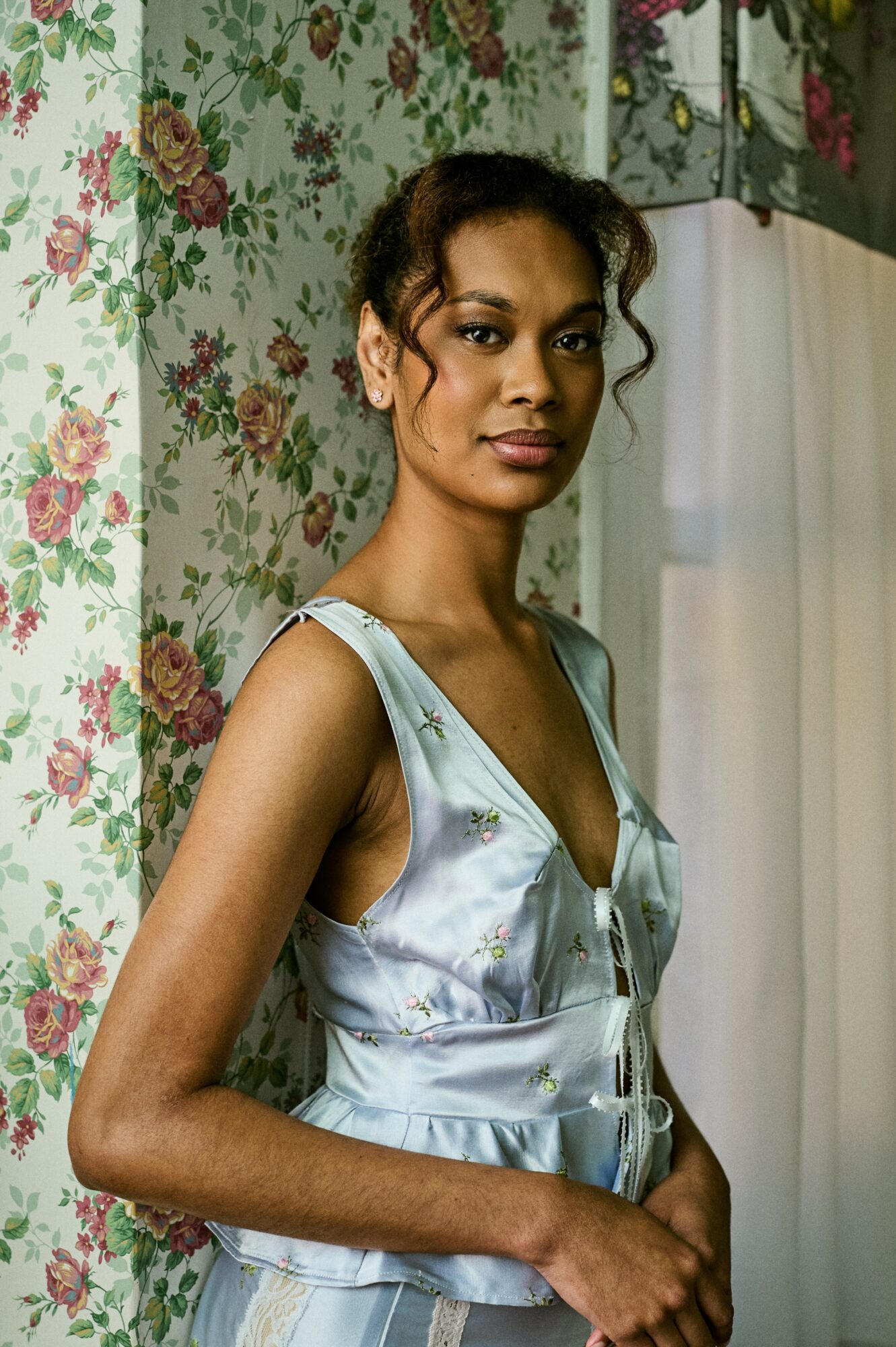 Young woman with dark hair in a sleeveless satin top standing in front of floral wallpaper and a curtain.
