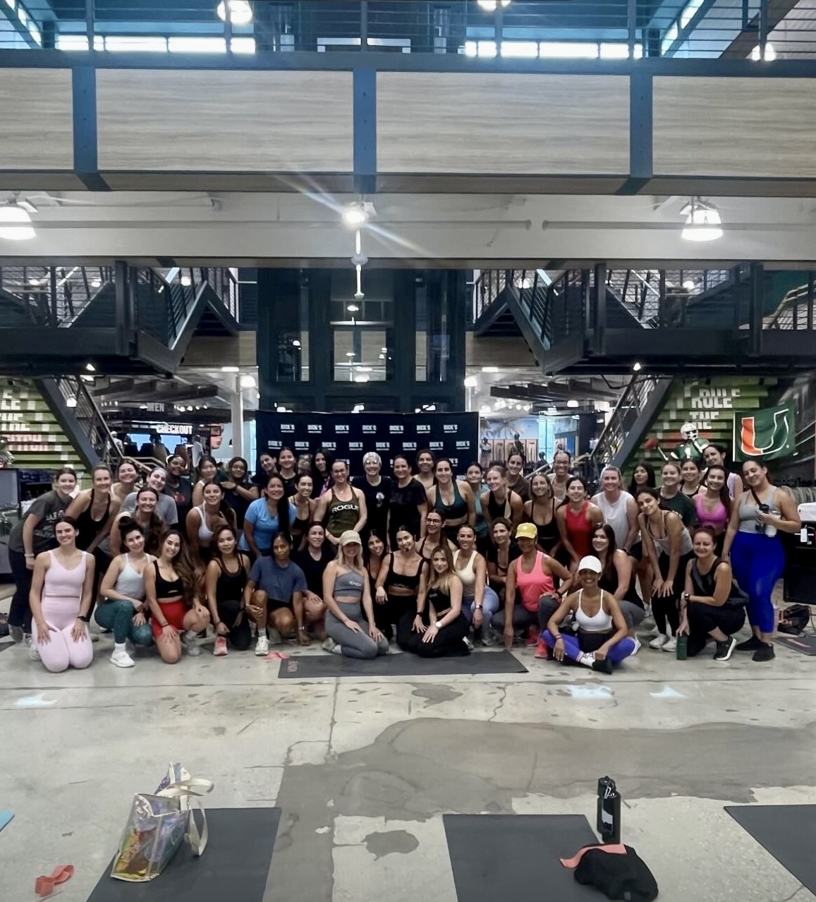 Group of people in a gym or fitness center, posing for a photo indoors, with equipment and stairs in the background.