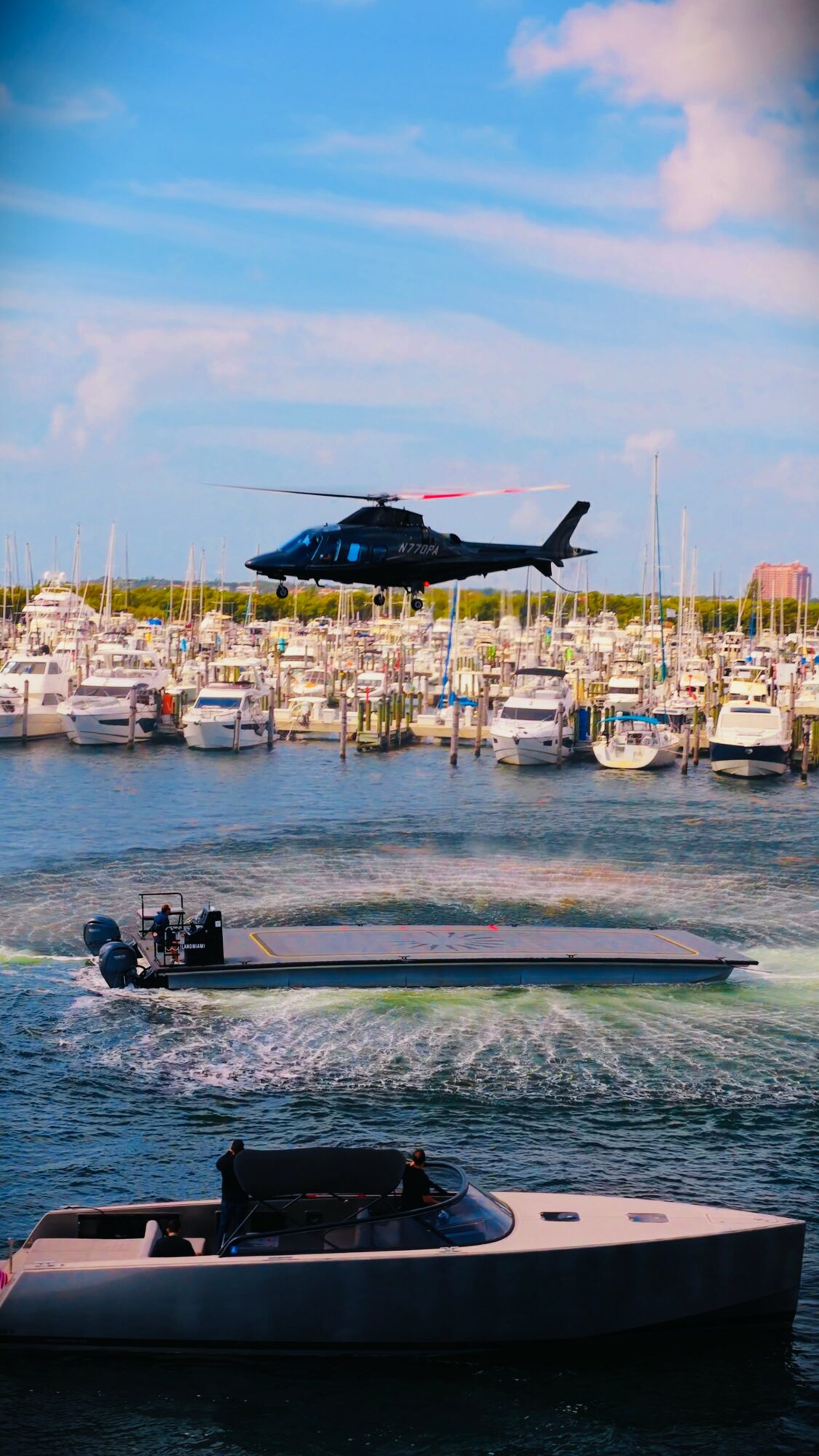 Black helicopter flying above a marina with boats, watercraft, and a dock, under a partly cloudy sky.