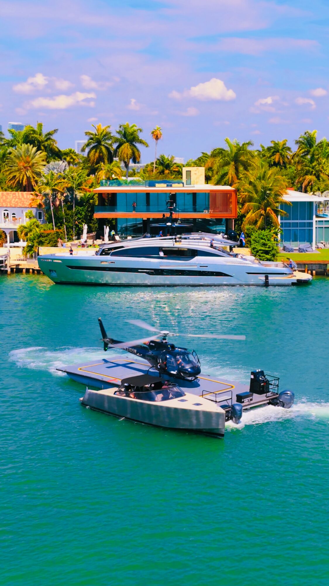 Two boats, a yacht and a smaller vessel, are on turquoise water with palm trees and colorful buildings in the background.