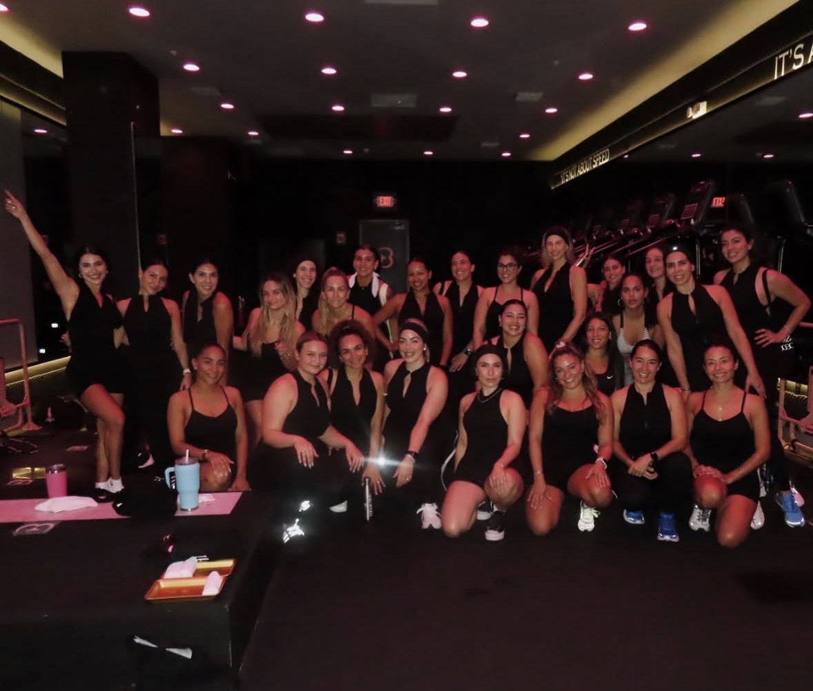 Group of women in black outfits posing in a dimly lit indoor space, some standing and some kneeling, with a dark background.