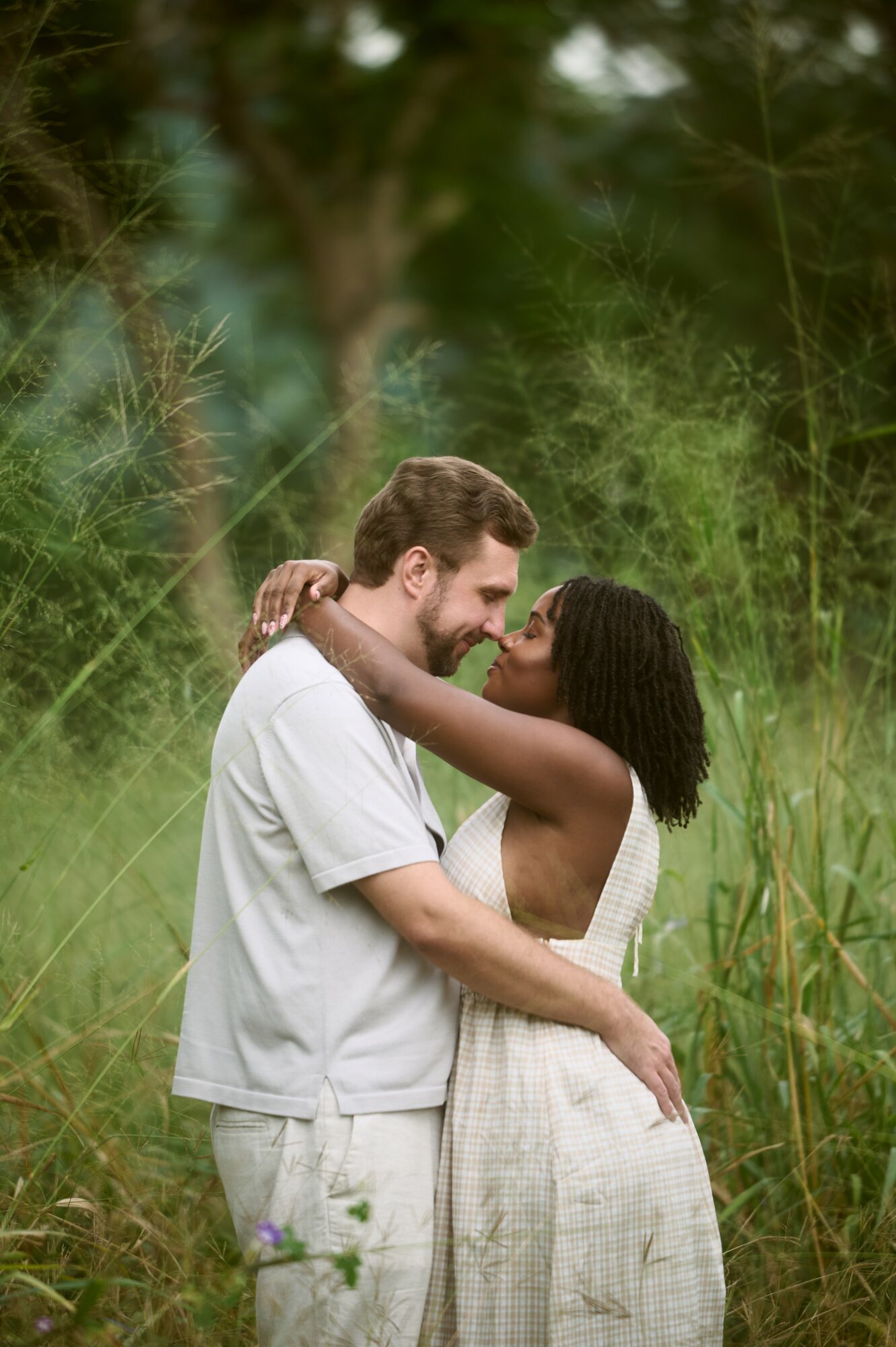 A couple embraces outdoors in a green natural setting, smiling and looking at each other.