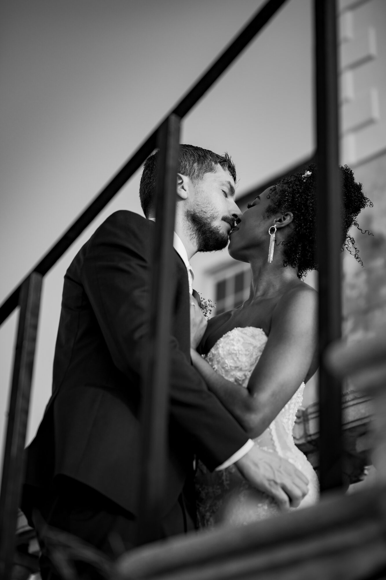 A couple in wedding attire sharing a kiss, seen through a metal railing, black and white photo.