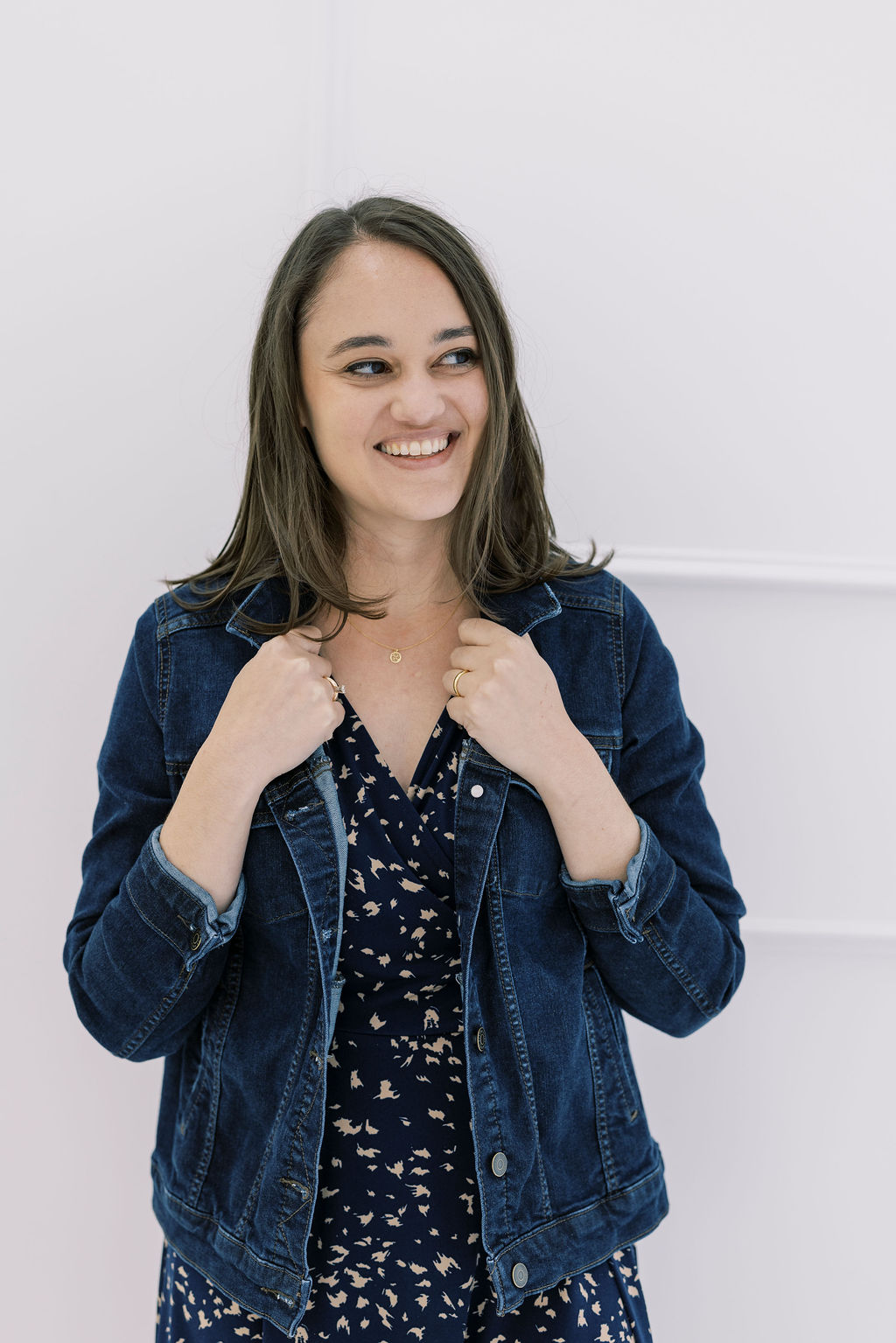 Young woman with shoulder-length dark hair smiling, wearing a denim jacket over a patterned top, standing against a plain white background.