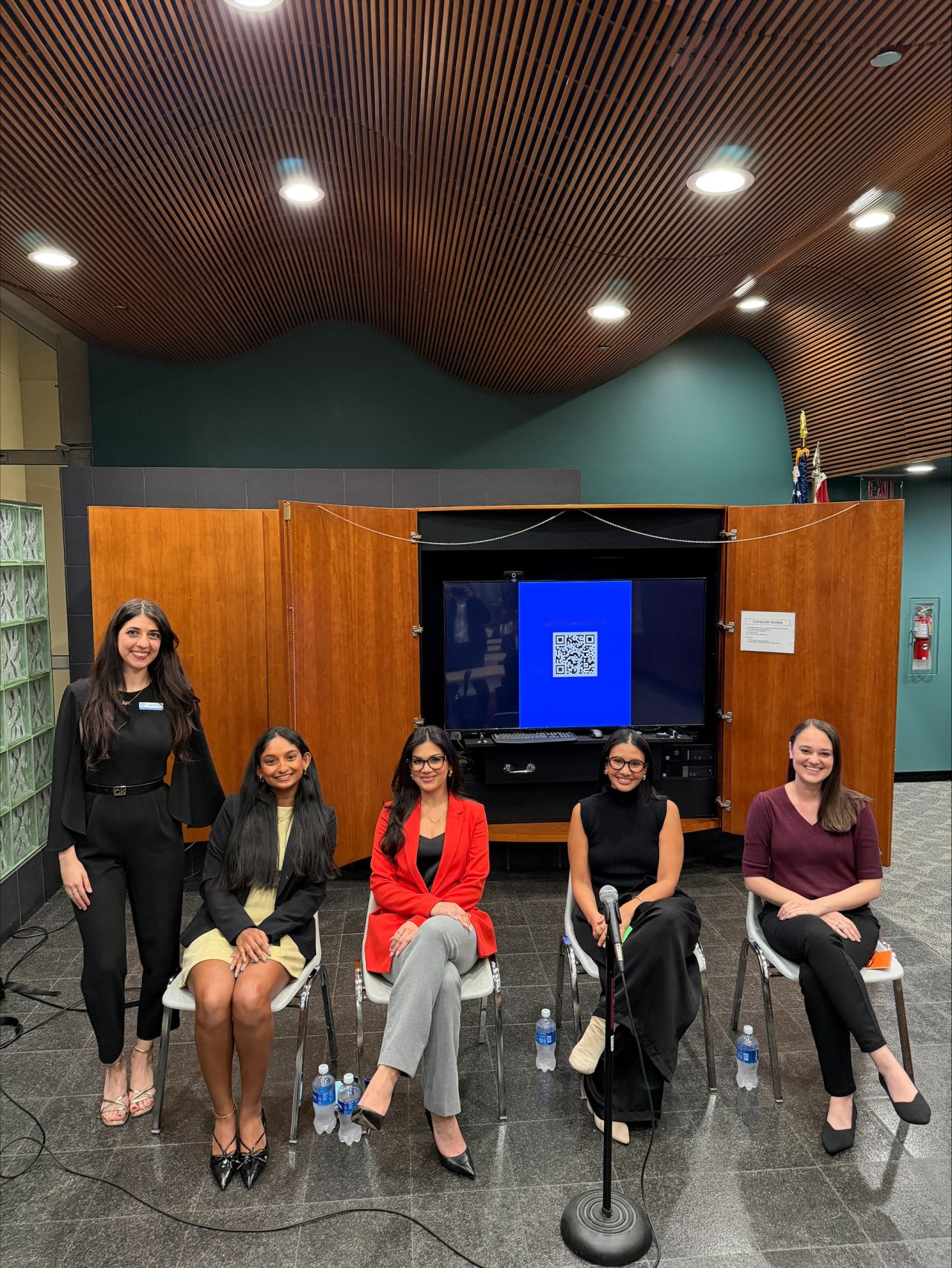 Five women sitting and standing in front of a large screen in a room with wooden ceiling panels.