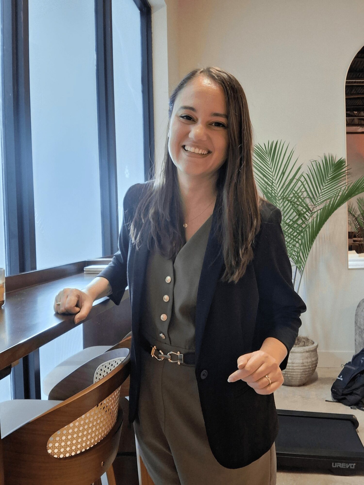 Smiling woman with long hair in a blazer and jumpsuit standing near a window with a plant behind her.