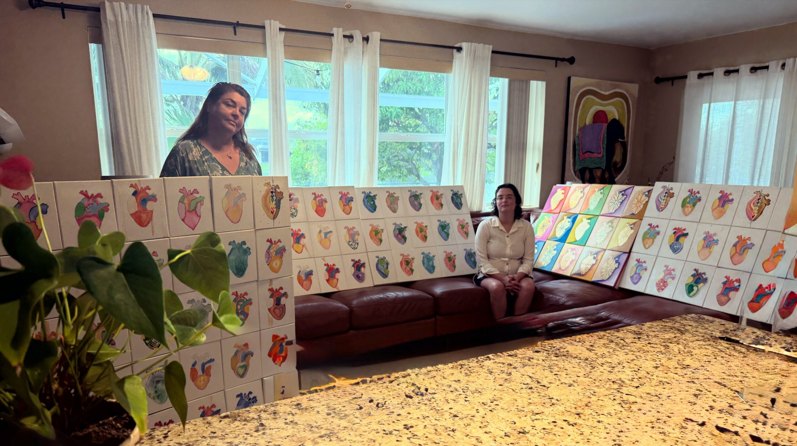 Two women in a room with large windows, surrounded by colorful artwork on display boards, and a patterned rug.