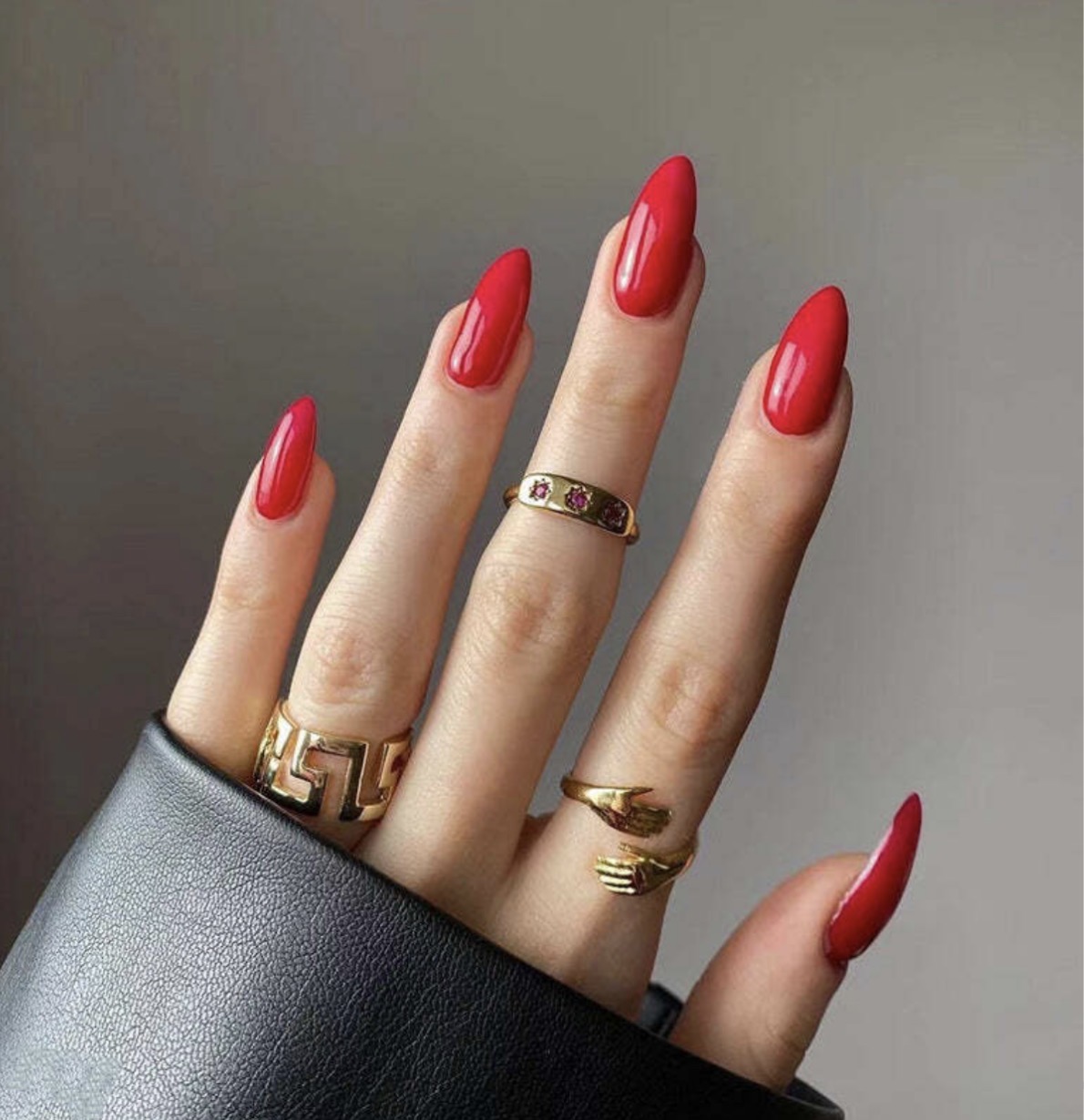 Hand with red manicured nails, wearing multiple gold rings, against a plain background.