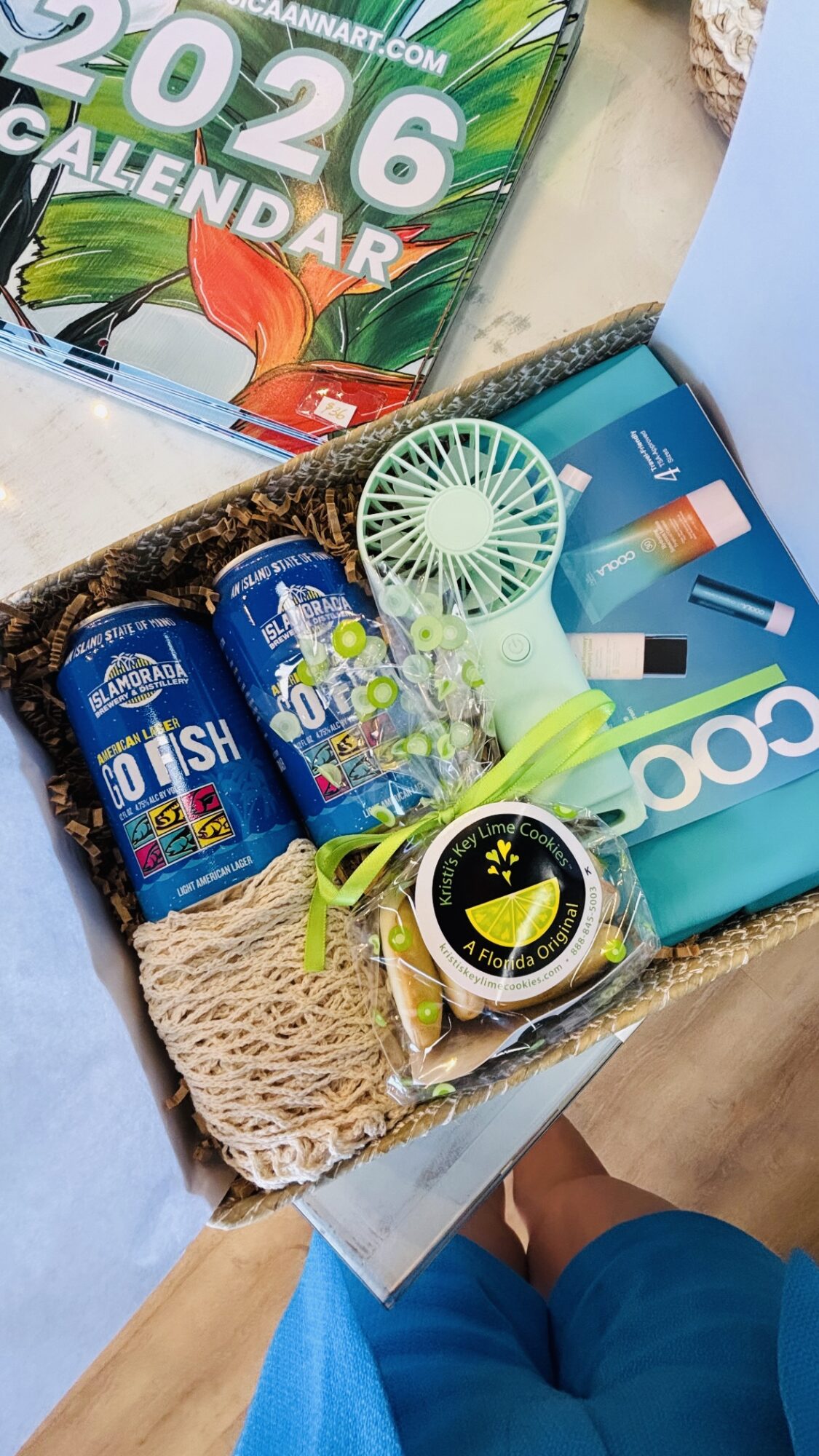 Open basket with sunscreen, a small fan, a watch, and a box of supplies, with a calendar in the background.