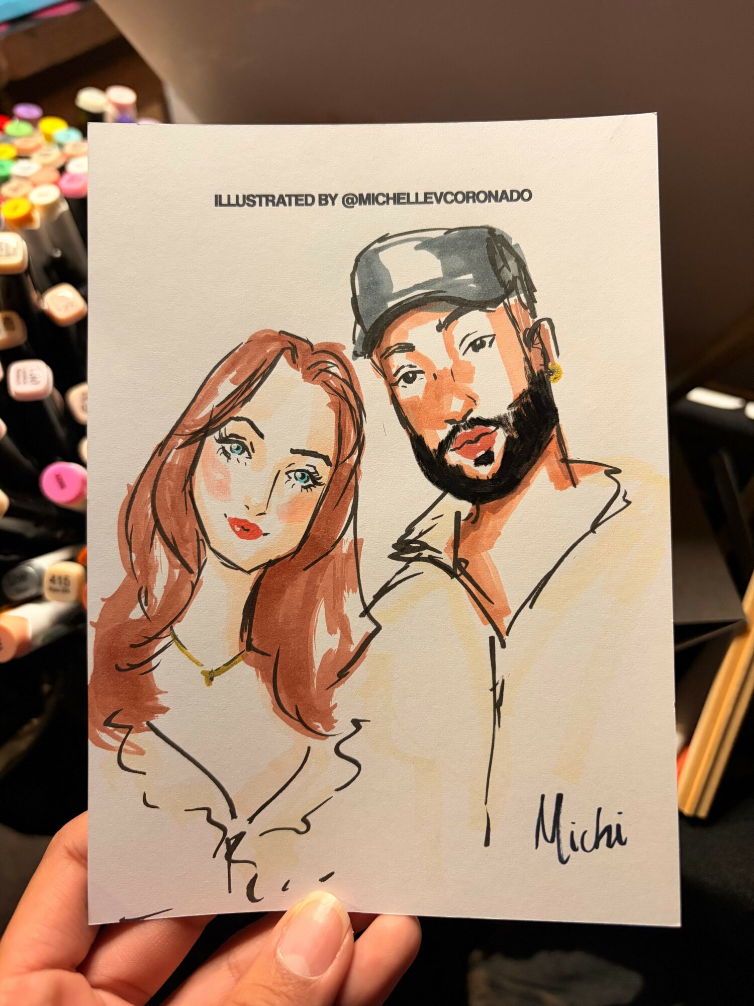 Hand holding a card with two illustrated people, a man with a beard and a woman with long hair, signatures on the side.