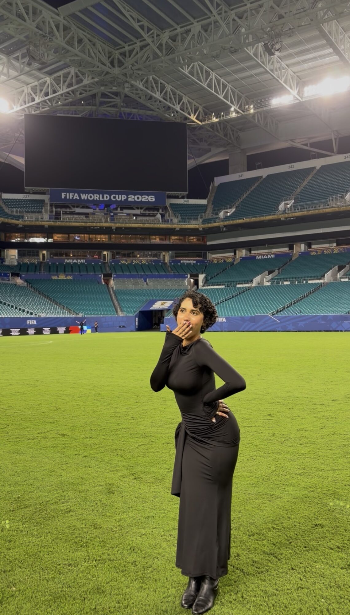 Woman in black dress standing on a soccer field inside a stadium, with empty seats and large screens in the background.