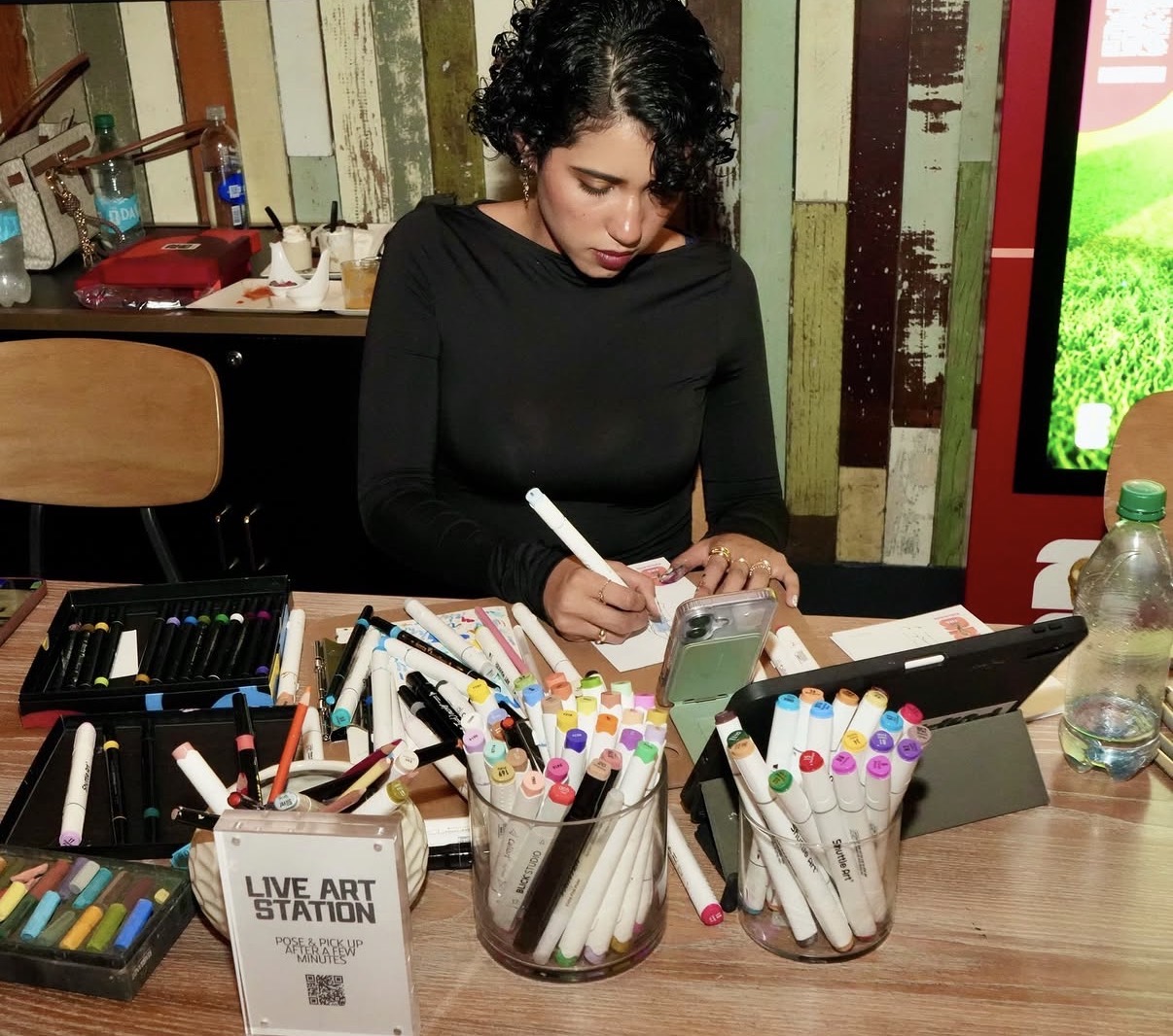 Woman with curly hair drawing with markers at a table filled with art supplies, in a colorful room.