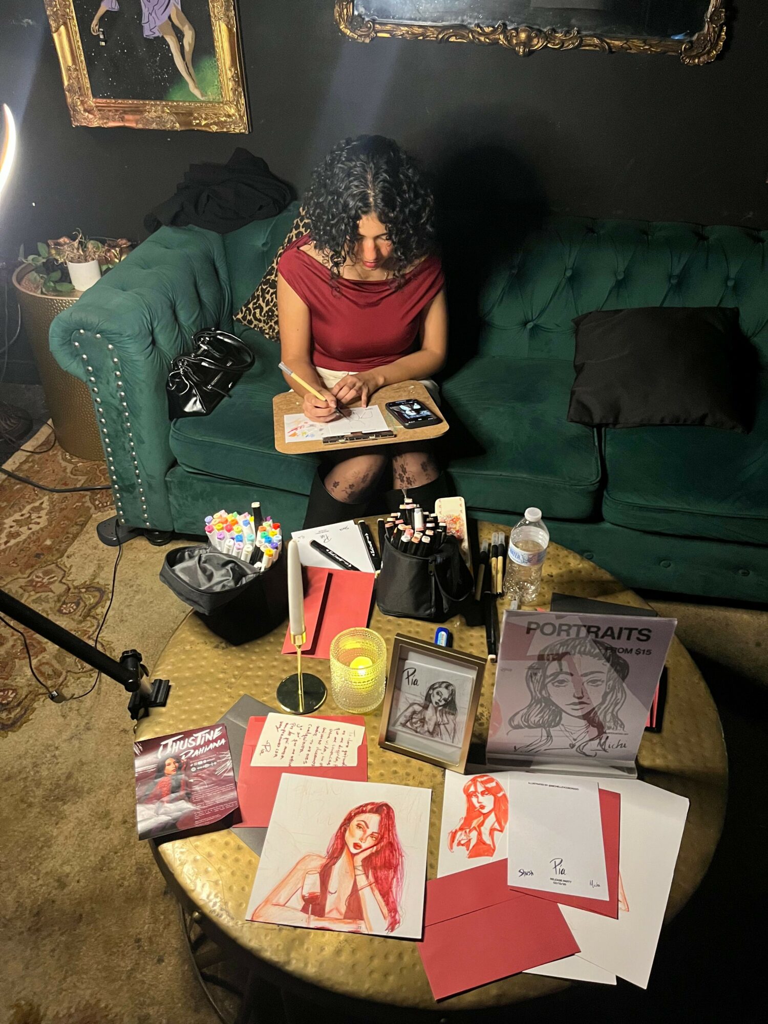 Woman with curly hair in red top sitting at table with art supplies, books, and framed pictures, in a room with green furniture.