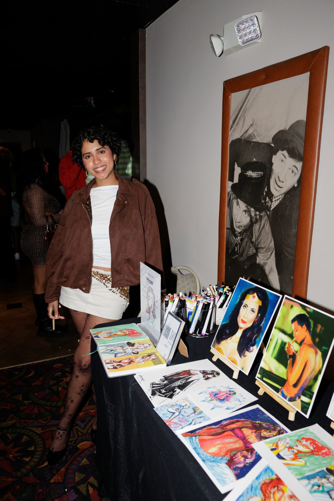 Woman with curly hair smiling standing next to a table with colorful artwork and photographs, framed black-and-white photo on wall behind her.