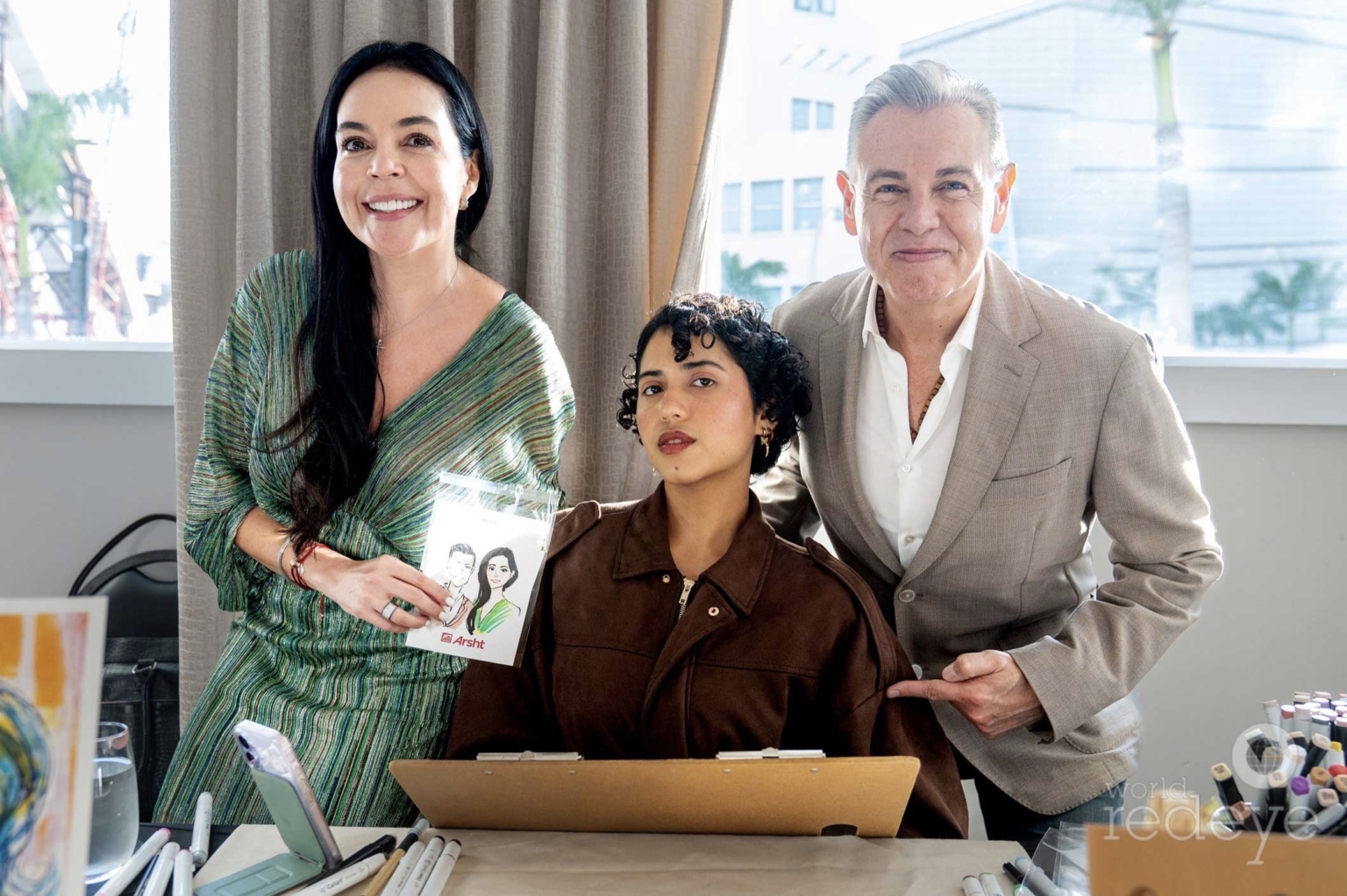 Three people, two women and one man, smiling indoors with a window in the background, one woman holding a photo, a laptop on a table.