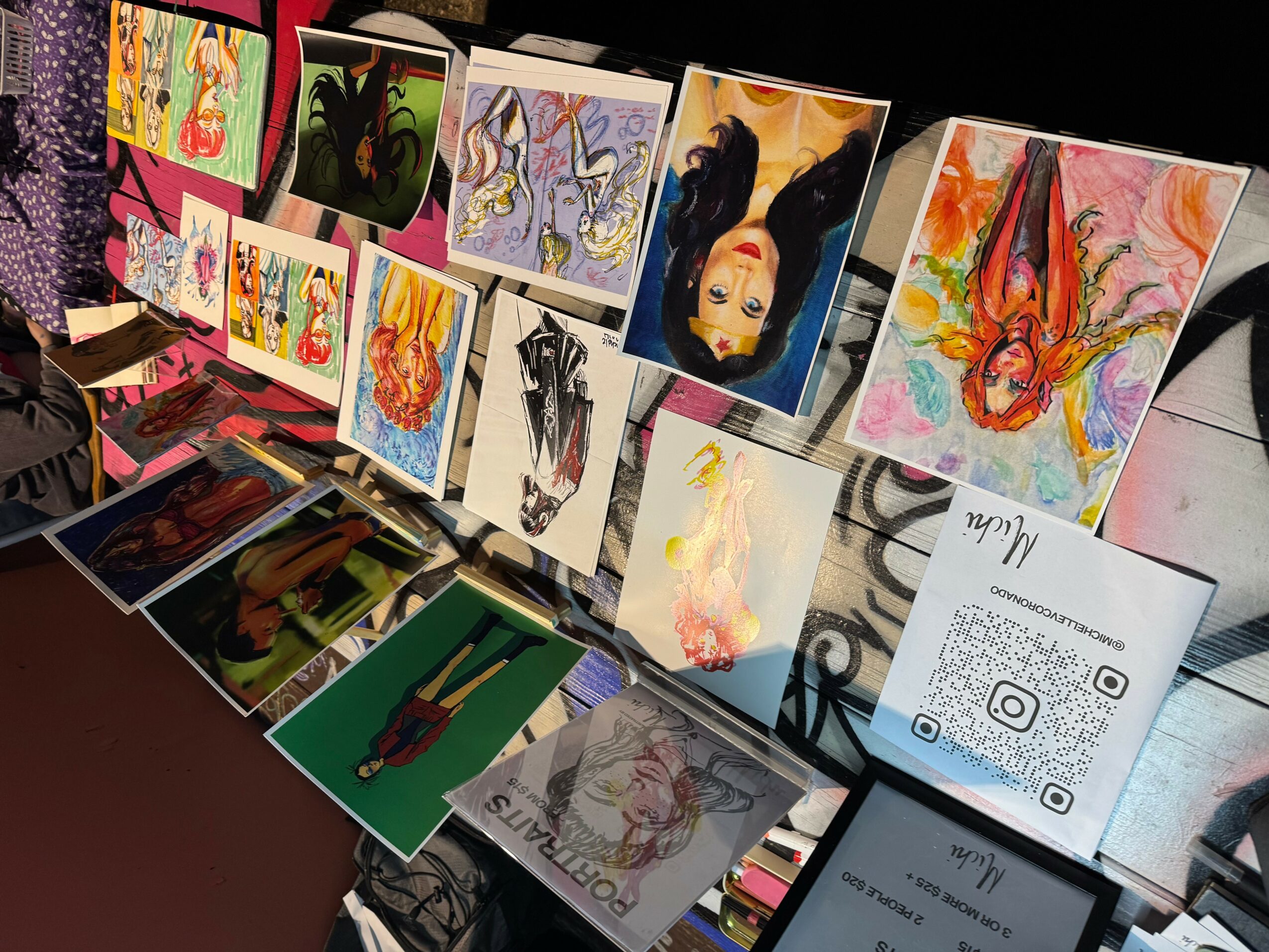 Display of colorful artwork and illustrations on a table, with a QR code sign in the foreground.