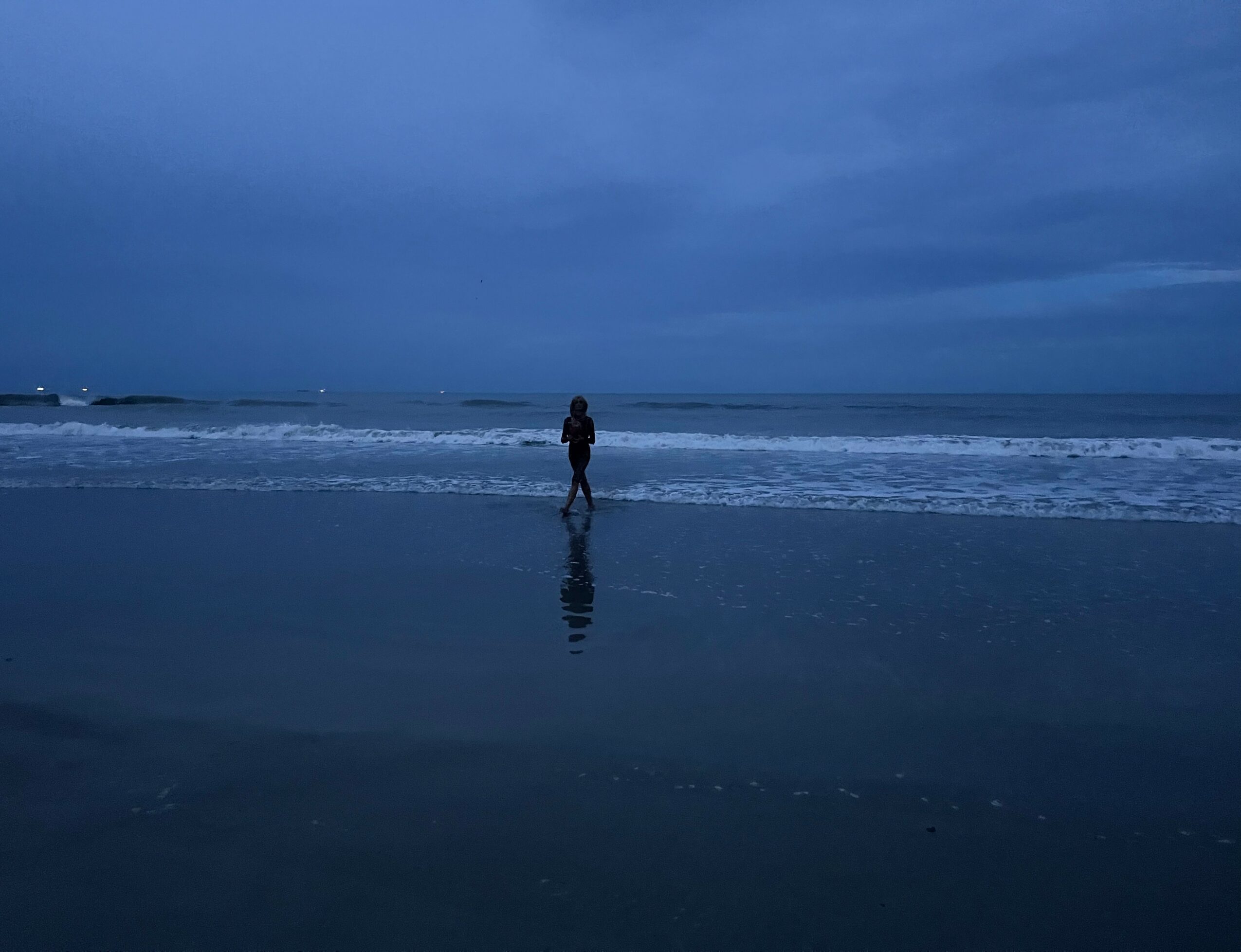 Person standing on the beach near the water, with a cloudy sky above, reflected in the wet sand.