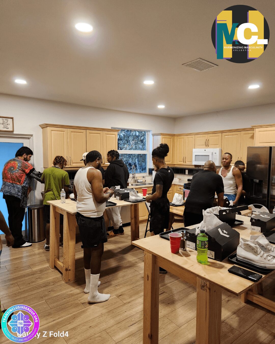Group of people in a kitchen with wooden cabinets and tables, some preparing food, others talking, with various items on the counters.