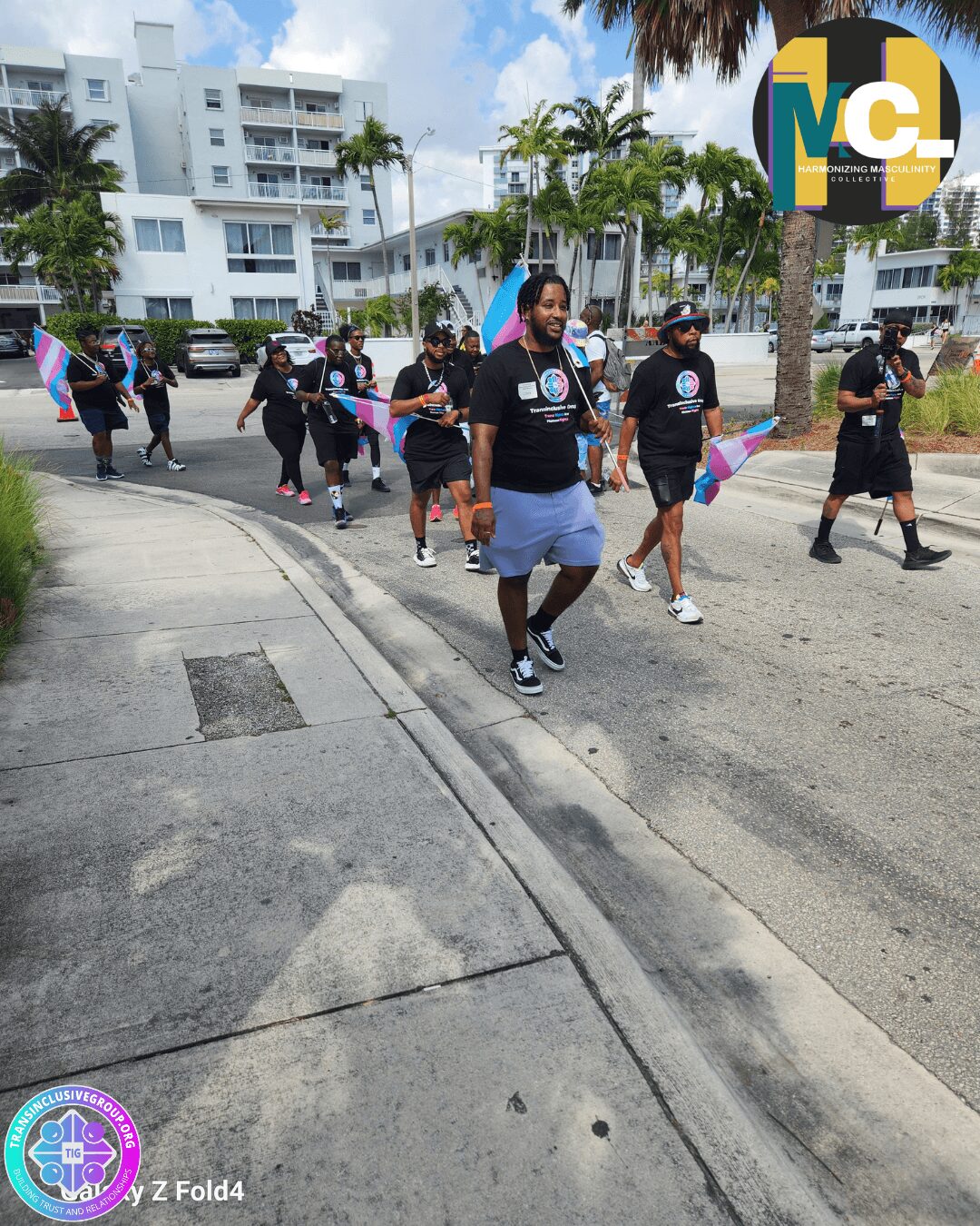 Group of people walking outdoors on a sunny day, some holding flags, with modern buildings and palm trees in background.