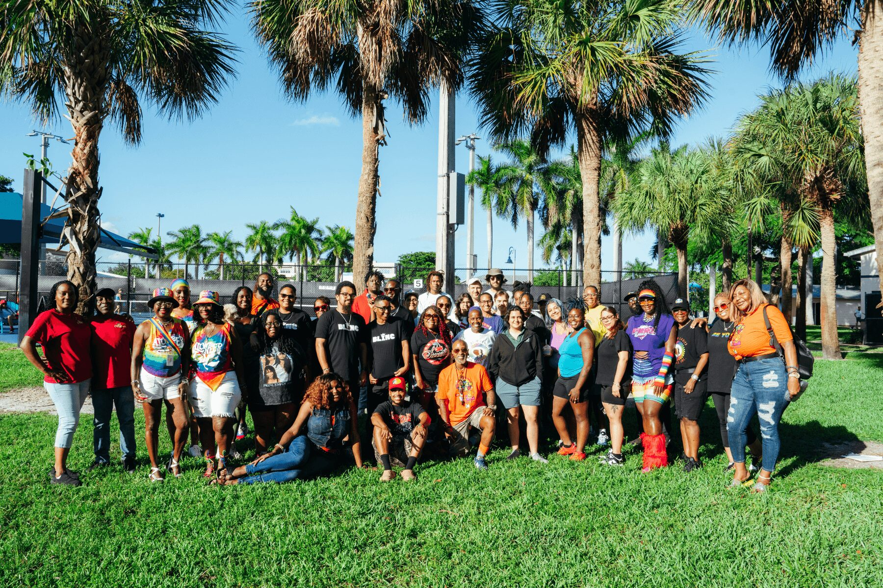 Group of people outdoors on grass with palm trees and blue sky in background, posing for a photo.