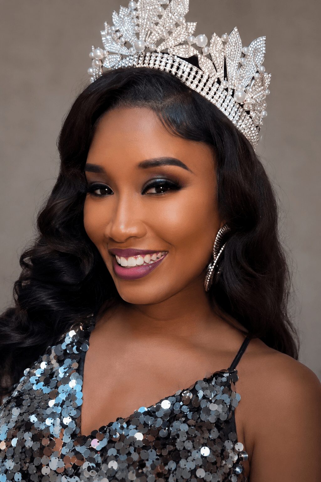 Woman wearing a crown and a sequined dress, smiling with dark wavy hair and earrings.