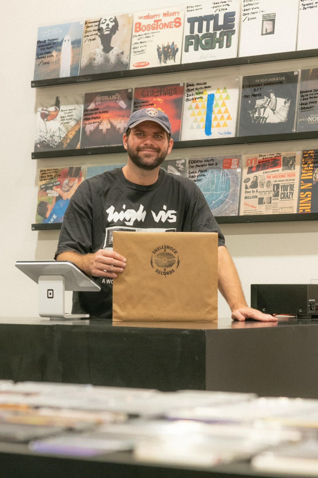 Person with a beard and cap standing behind a counter with a paper bag, shelves with books or records in background.