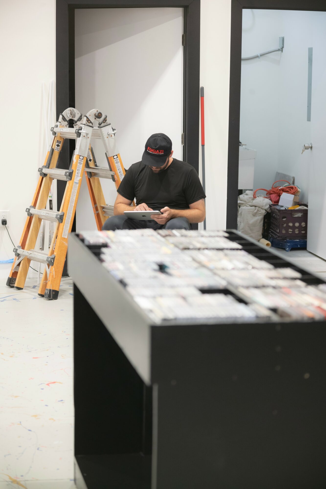 Person sitting on the floor using a phone, with a ladder and storage area nearby, in a modern interior space.