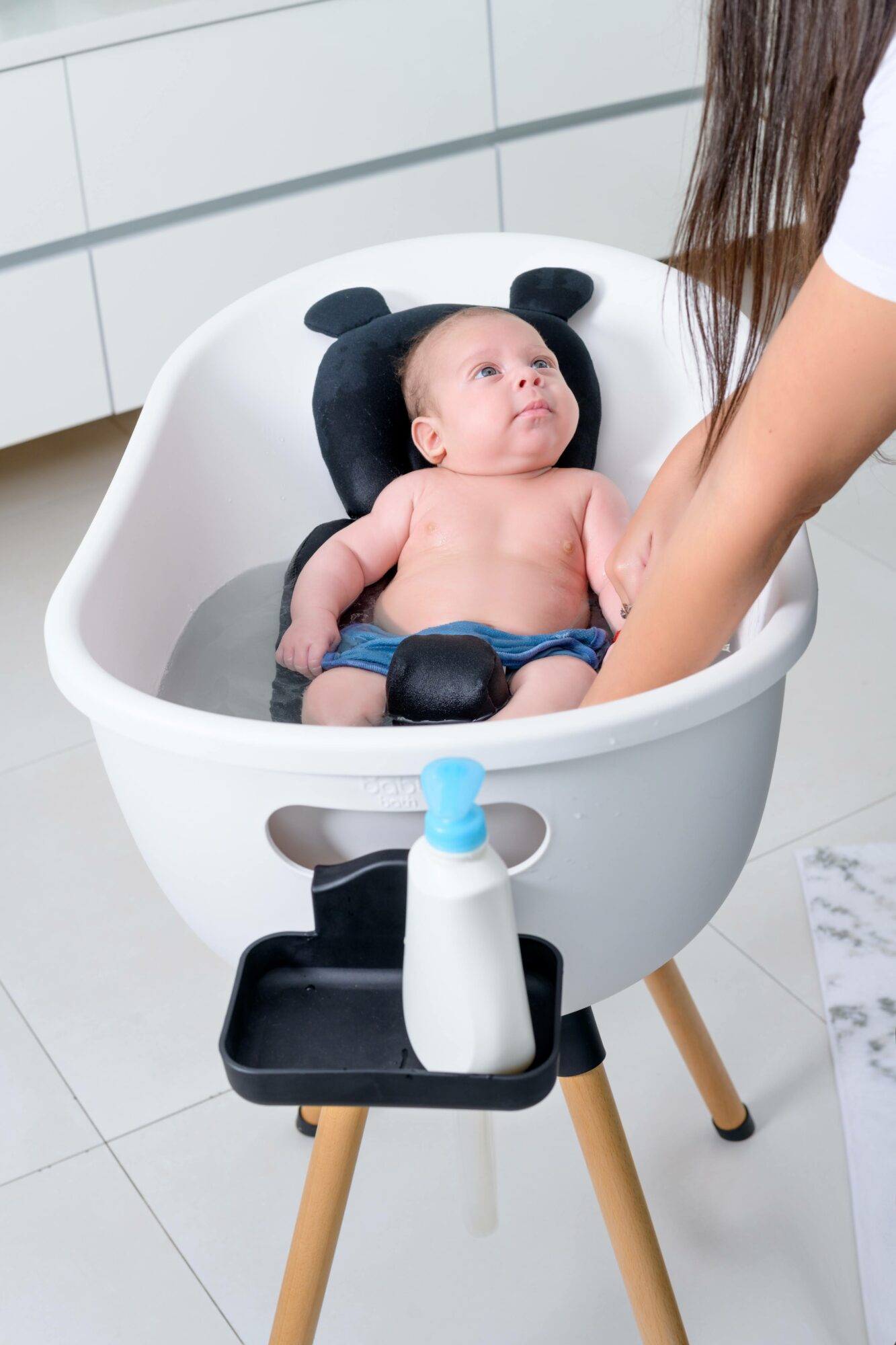 Baby lying in a white baby bath with a black headrest, looking up at a person washing them, in a bright room.