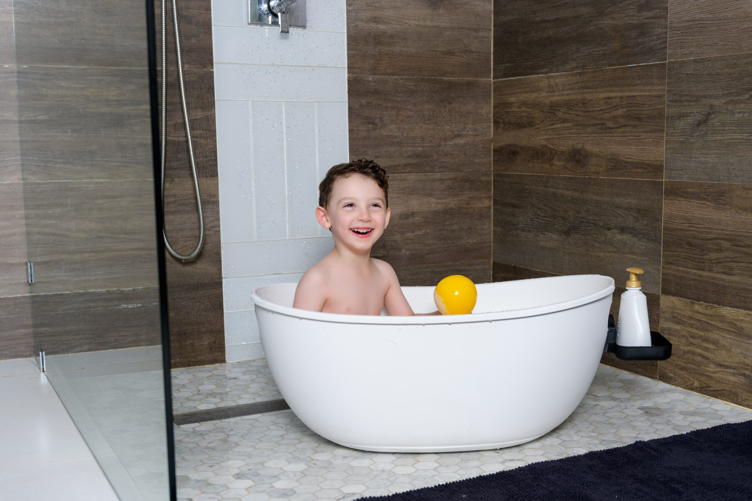 Child smiling in a white bathtub with a yellow ball, in a modern bathroom with wood and tile walls.