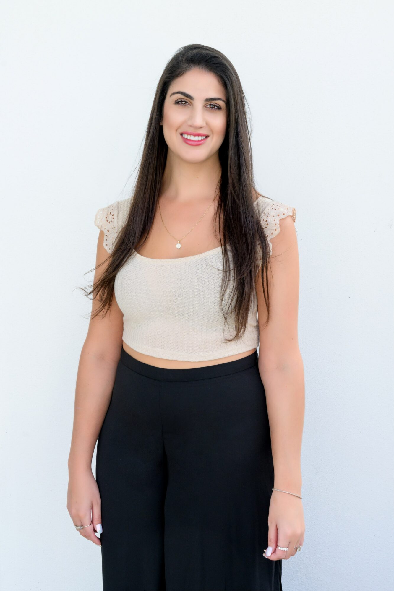 Young woman with long dark hair smiling, wearing a beige top and black pants, standing against a white background.