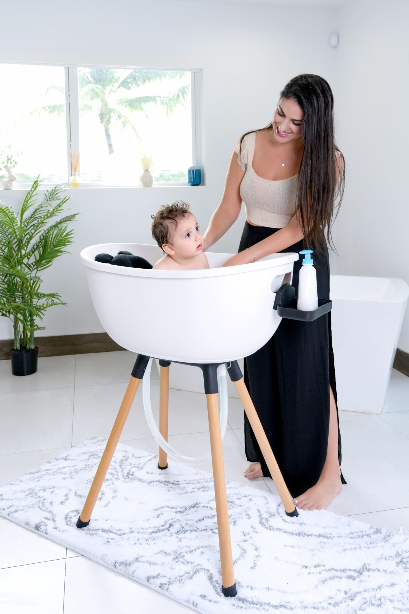 Woman with long dark hair smiling at a baby in a white bathtub on wooden legs in a bright room with a plant and window.