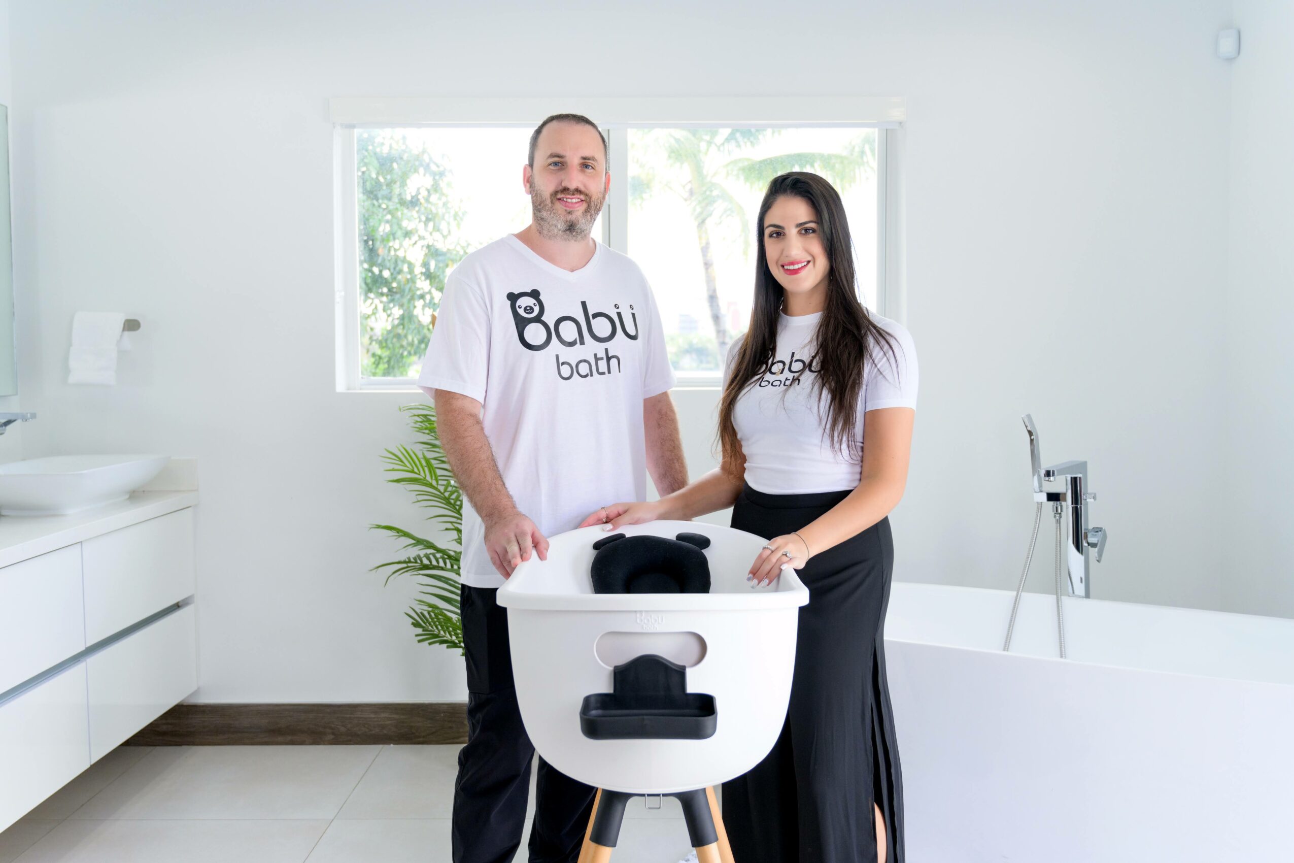 Two people standing in a bright bathroom with a baby bathtub, smiling at the camera.