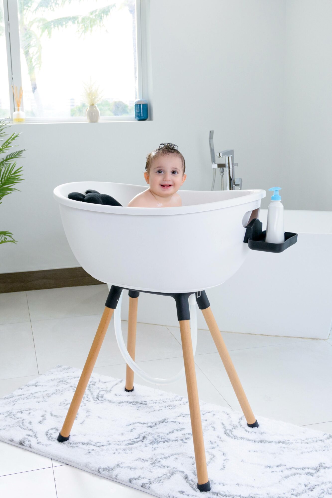 Baby in a white bathtub on wooden legs, smiling, in a bright bathroom with a window and a plant.