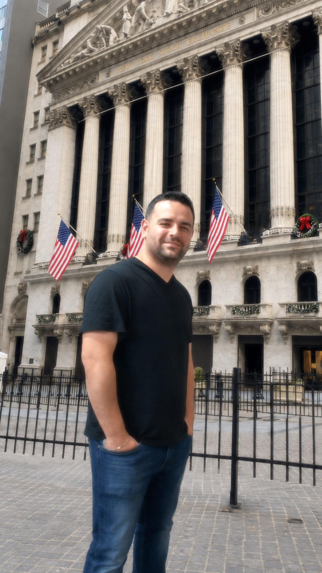 Man standing in front of a historic building with tall columns and American flags, smiling, wearing a black shirt and jeans.