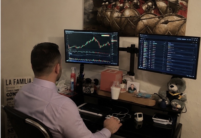Person with short hair in a pink shirt working at a desk with two monitors displaying stock charts and data.