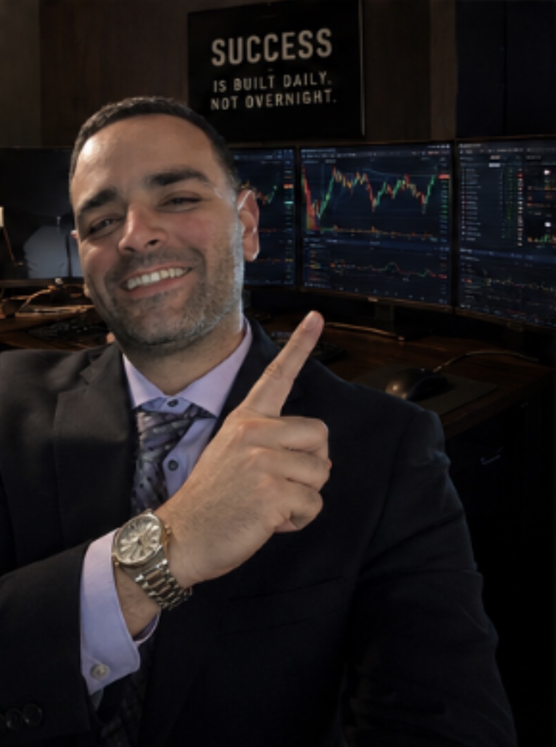 Man in suit smiling and pointing upward, with multiple computer monitors displaying financial charts behind him.