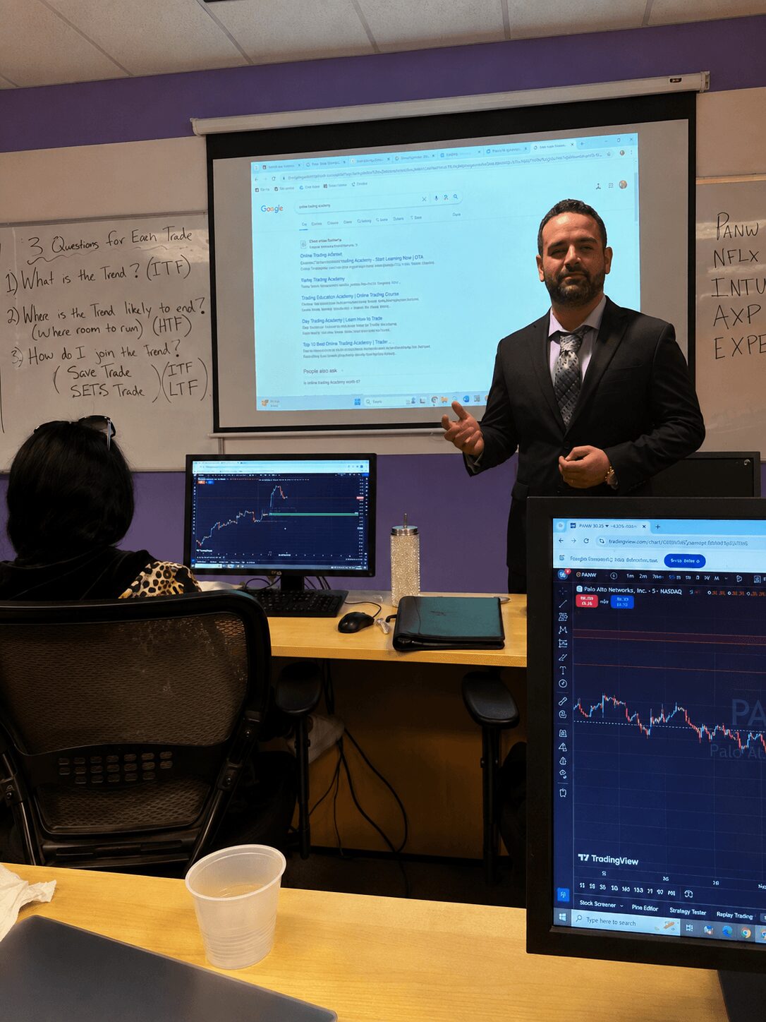 Man in suit standing in front of a classroom with a presentation screen, two computer monitors showing graphs, and a whiteboard.