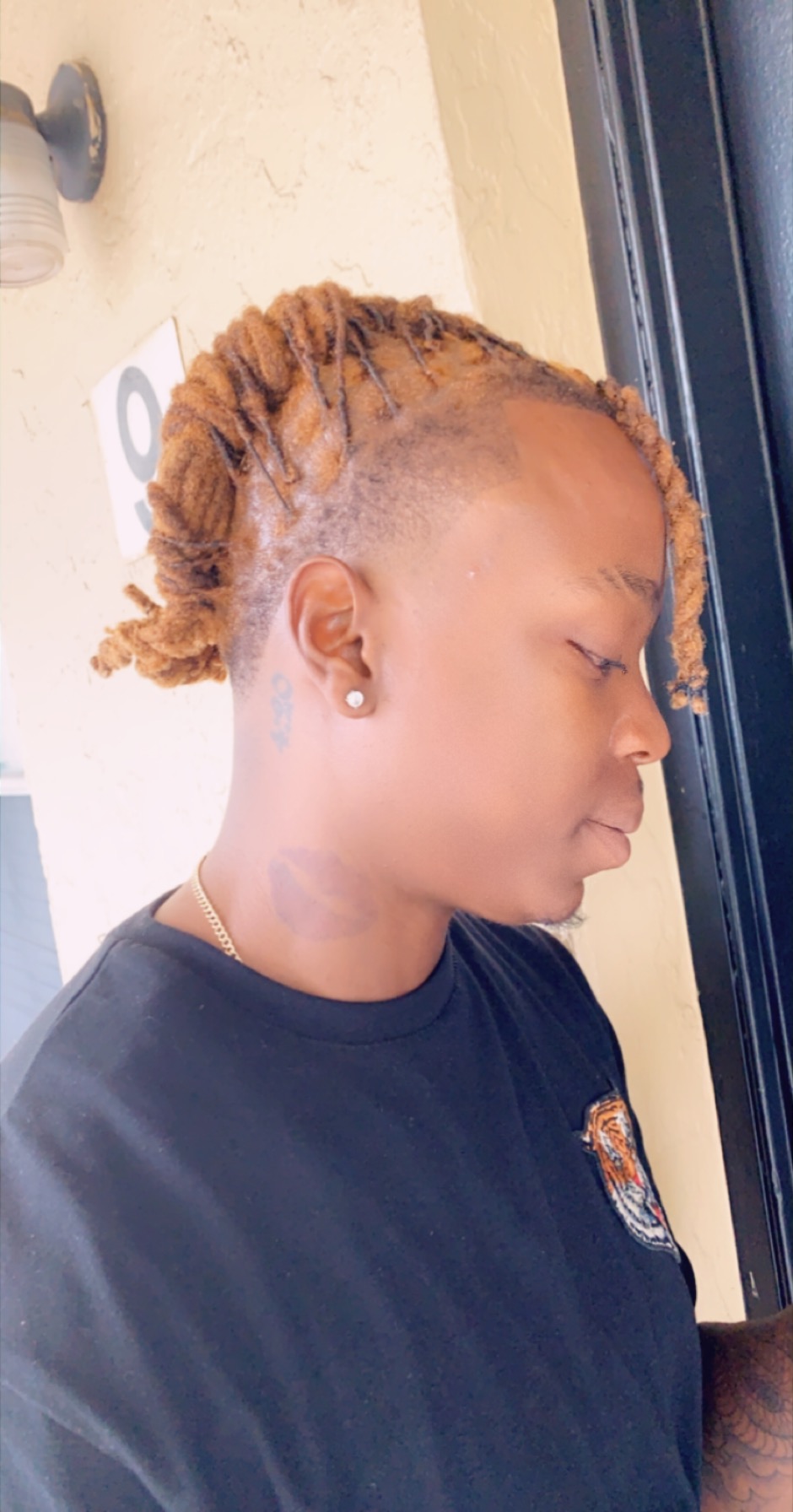 Side profile of a person with styled dreadlocks wearing a dark shirt, standing near a window.