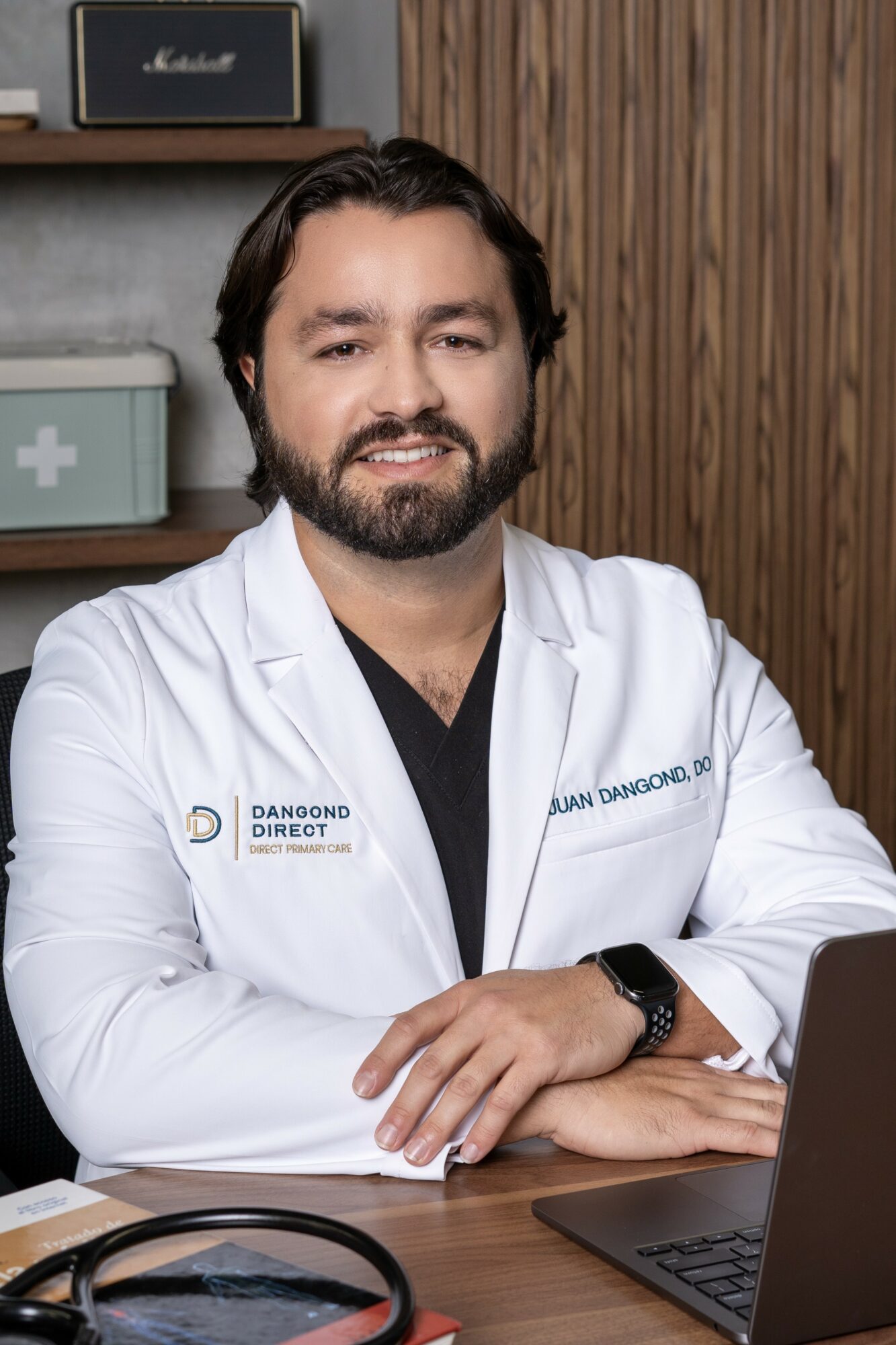 Male doctor with beard and dark hair, wearing a white coat, sitting at a desk with a laptop, wooden wall background.