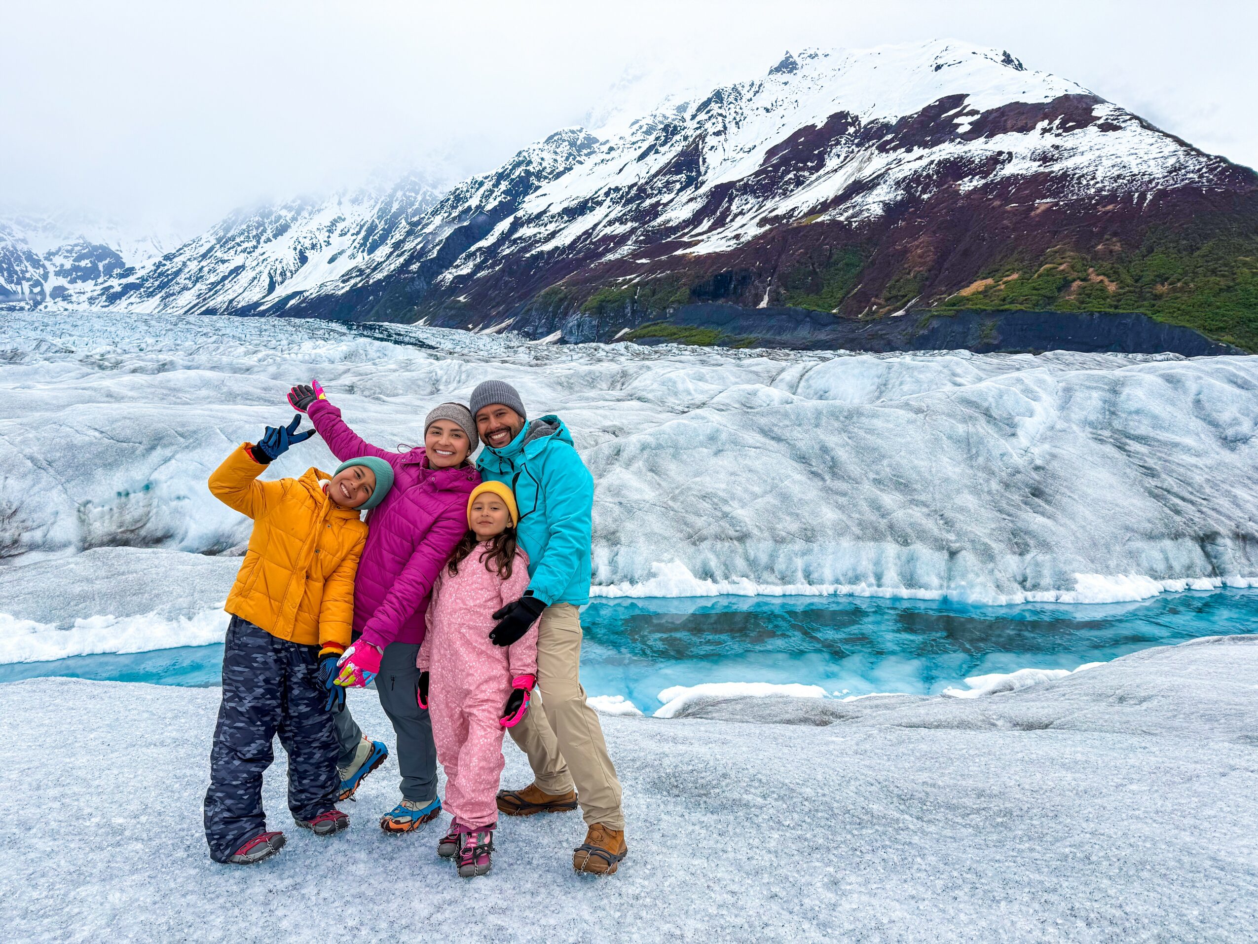 Group of four children in colorful winter clothing standing on icy landscape with snow-covered mountains and glacier in background.