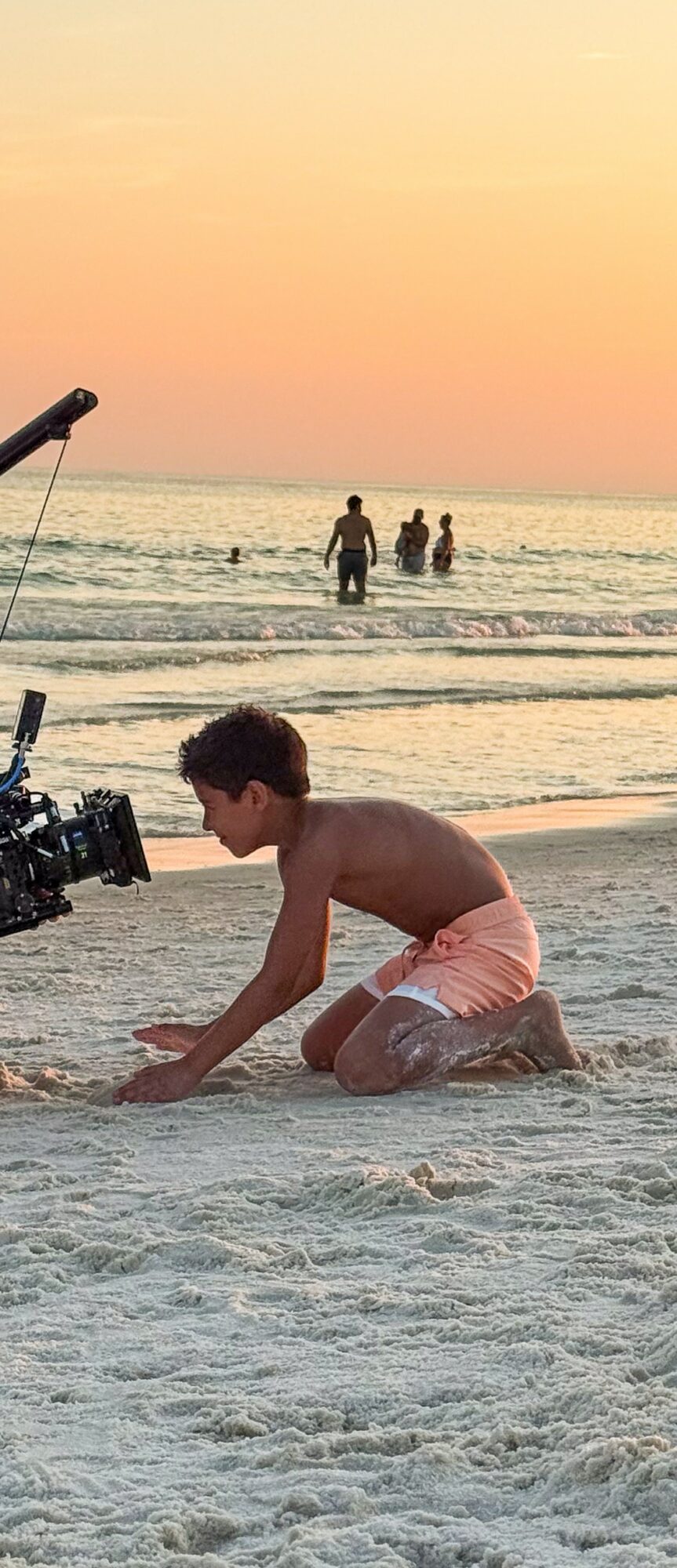 Child kneeling on sand at the beach during sunset, with people in the water and a camera on a tripod nearby.