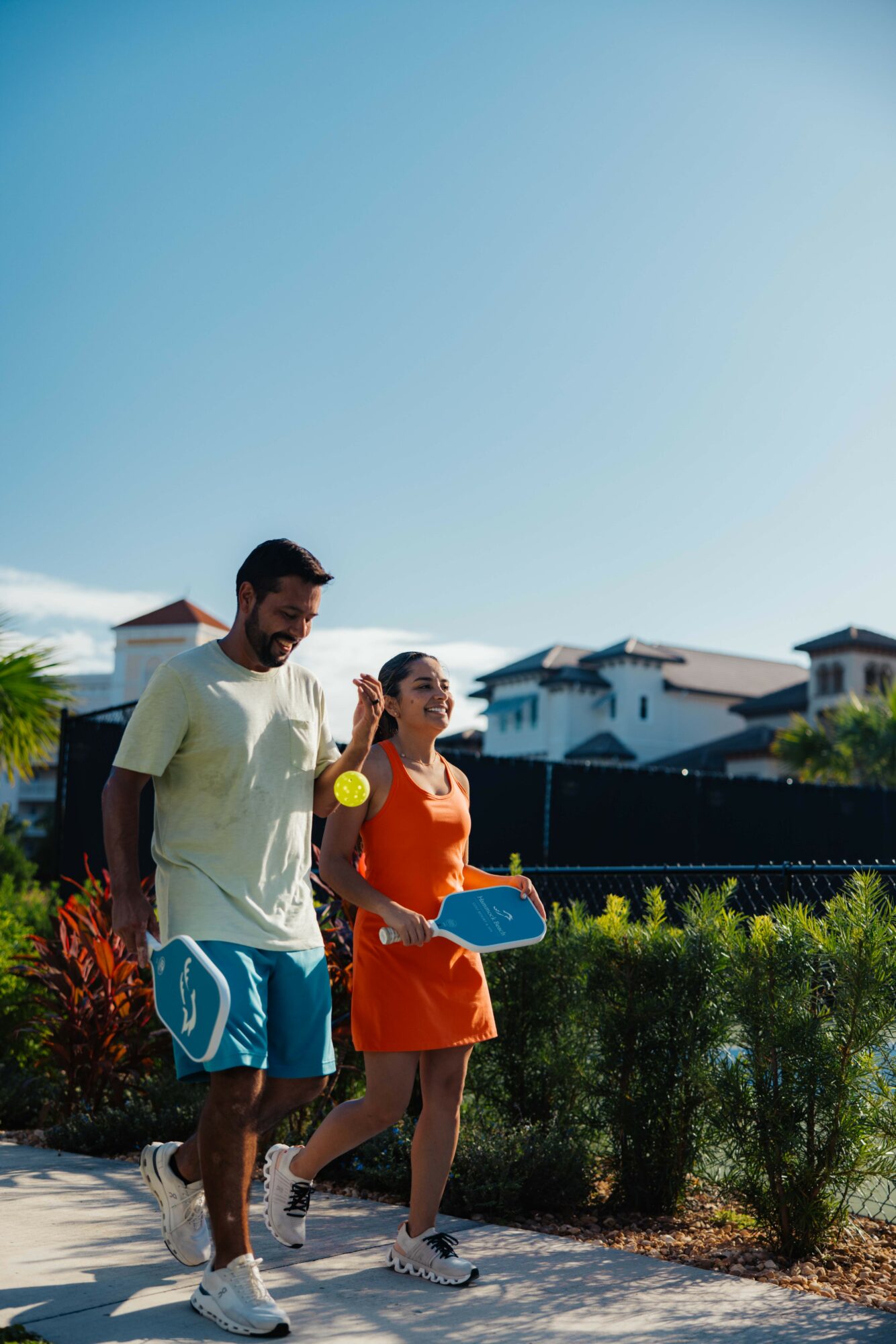 Two people walking outdoors on a sunny day, with buildings and greenery in the background.