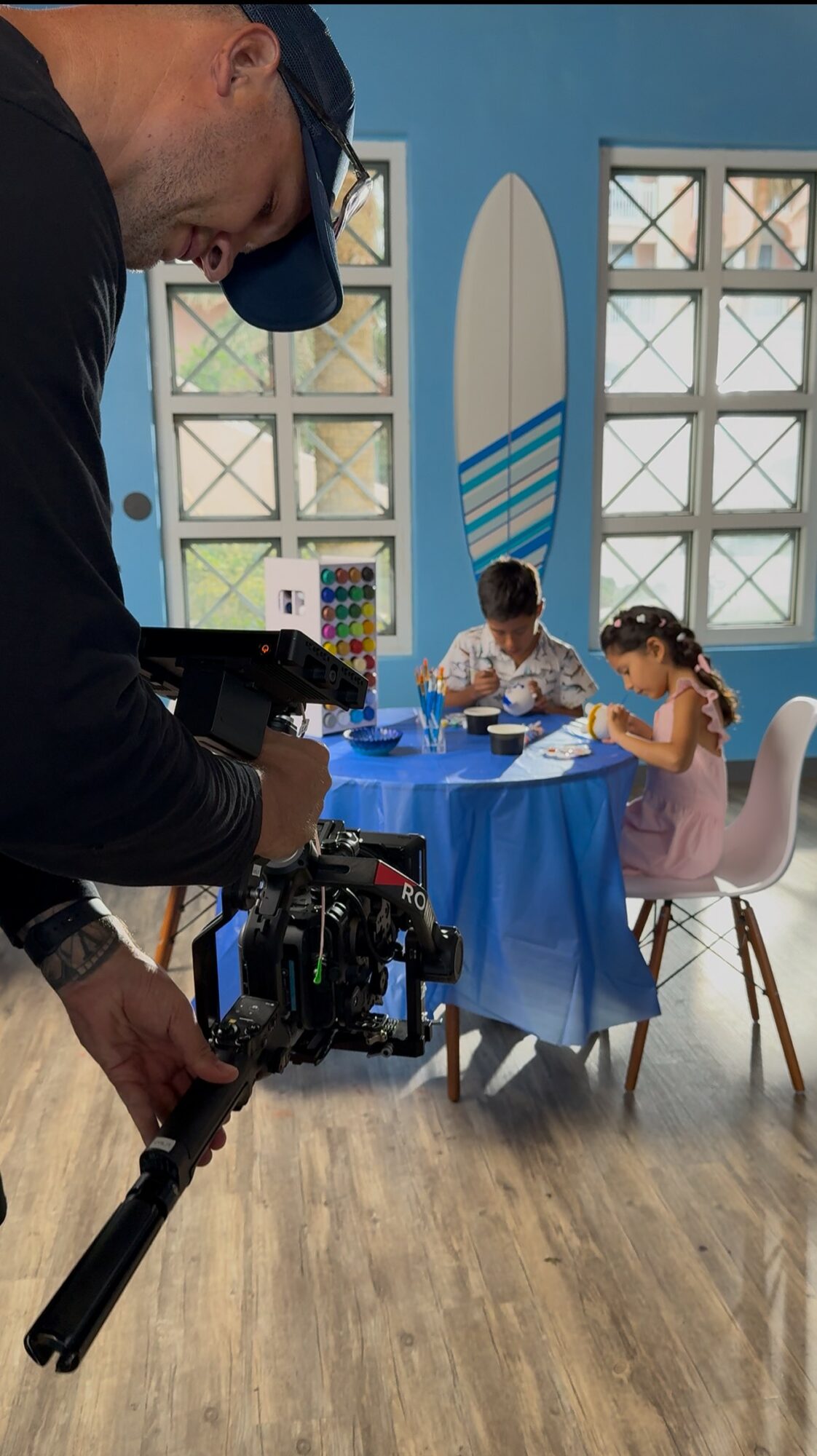 Person filming two children sitting at a table with a blue tablecloth in a brightly lit room with large windows and surfboard decor.