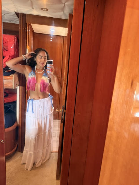 Young woman with long hair in a colorful top and white skirt taking a mirror selfie in a wooden-paneled room.