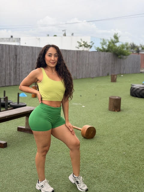 Young woman with long curly hair in a yellow crop top and green shorts outdoors on grass, holding a wooden mallet.