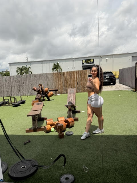 Woman in workout clothes taking a selfie outdoors with exercise equipment and a person in the background.