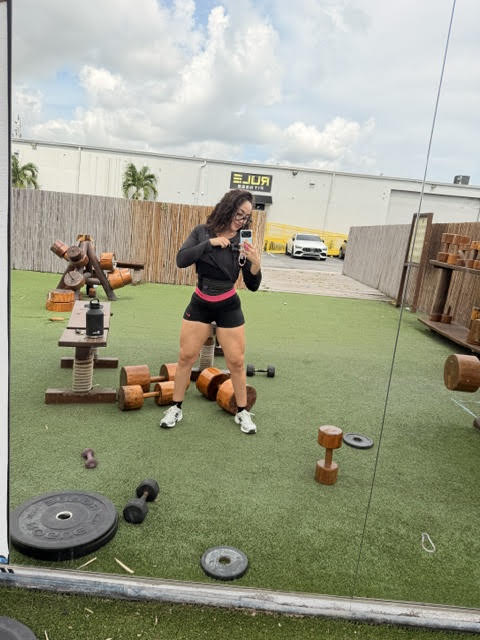 Person taking a mirror selfie outdoors with gym equipment and a fence in the background.