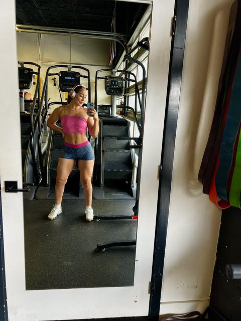 Woman taking a mirror selfie in a gym, wearing a pink top and shorts, with gym equipment in the background.